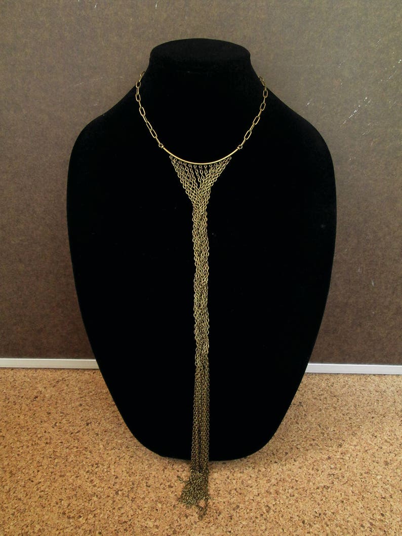 Puede incluir: Un collar largo de cadena dorada con una parte superior curva y un borla de cadenas que cuelga.