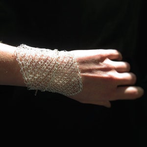 May include: A silver wire mesh bracelet with a delicate, intricate design. The bracelet is worn on a person's wrist.