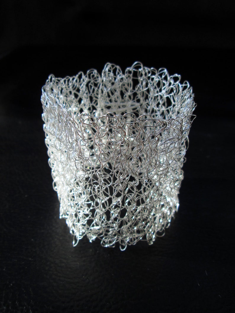 May include: A silver wire bracelet with a delicate, crocheted design. The bracelet is made of thin, interwoven wire that creates a lacy, openwork pattern.