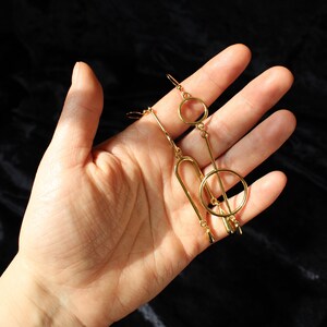 Op de afbeelding: Een paar gouden geometrische oorbellen met een cirkel, een lange staaf en een gebogen vorm. De oorbellen hangen en hebben een druppelvormig detail aan de onderkant.