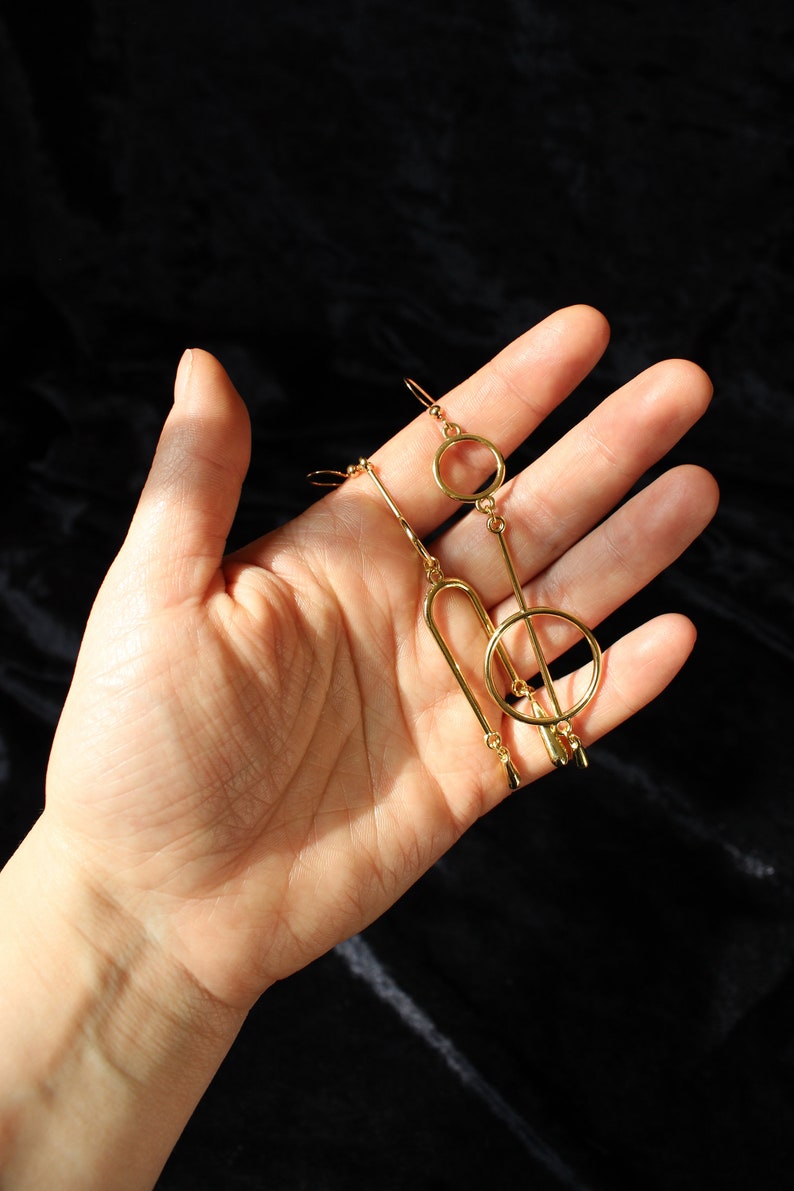Op de afbeelding: Een paar gouden geometrische oorbellen met een cirkel, een lange staaf en een gebogen vorm. De oorbellen hangen en hebben een druppelvormig detail aan de onderkant.