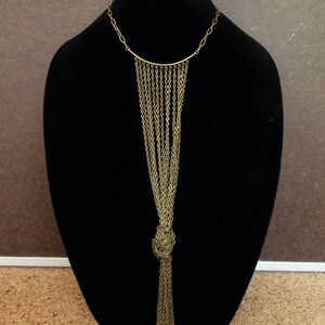 Puede incluir: Un collar de cadena dorada con un largo fleco anudado. El collar se muestra en un busto de maniqu&iacute; negro.