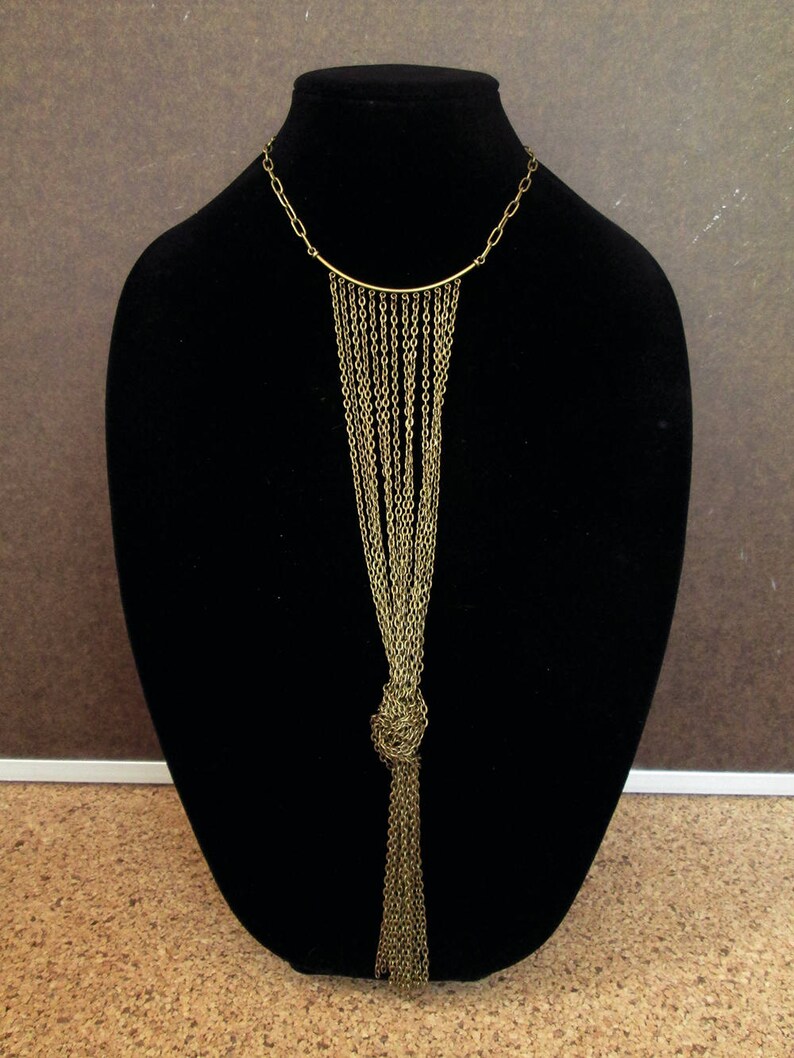 Puede incluir: Un collar de cadena dorada con un largo fleco anudado. El collar se muestra en un busto de maniqu&iacute; negro.