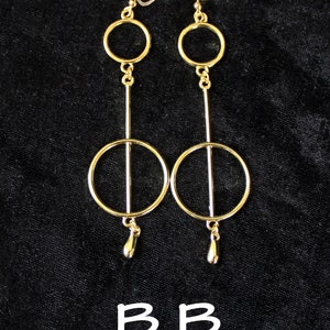 Op de afbeelding: Een paar gouden oorbellen met twee cirkels en een traanvormige hanger. De oorbellen zijn op een zwarte achtergrond.