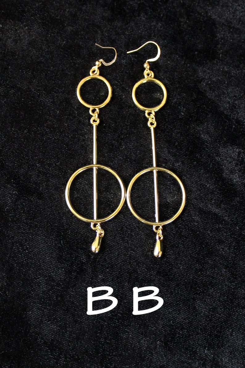 Op de afbeelding: Een paar gouden oorbellen met twee cirkels en een traanvormige hanger. De oorbellen zijn op een zwarte achtergrond.
