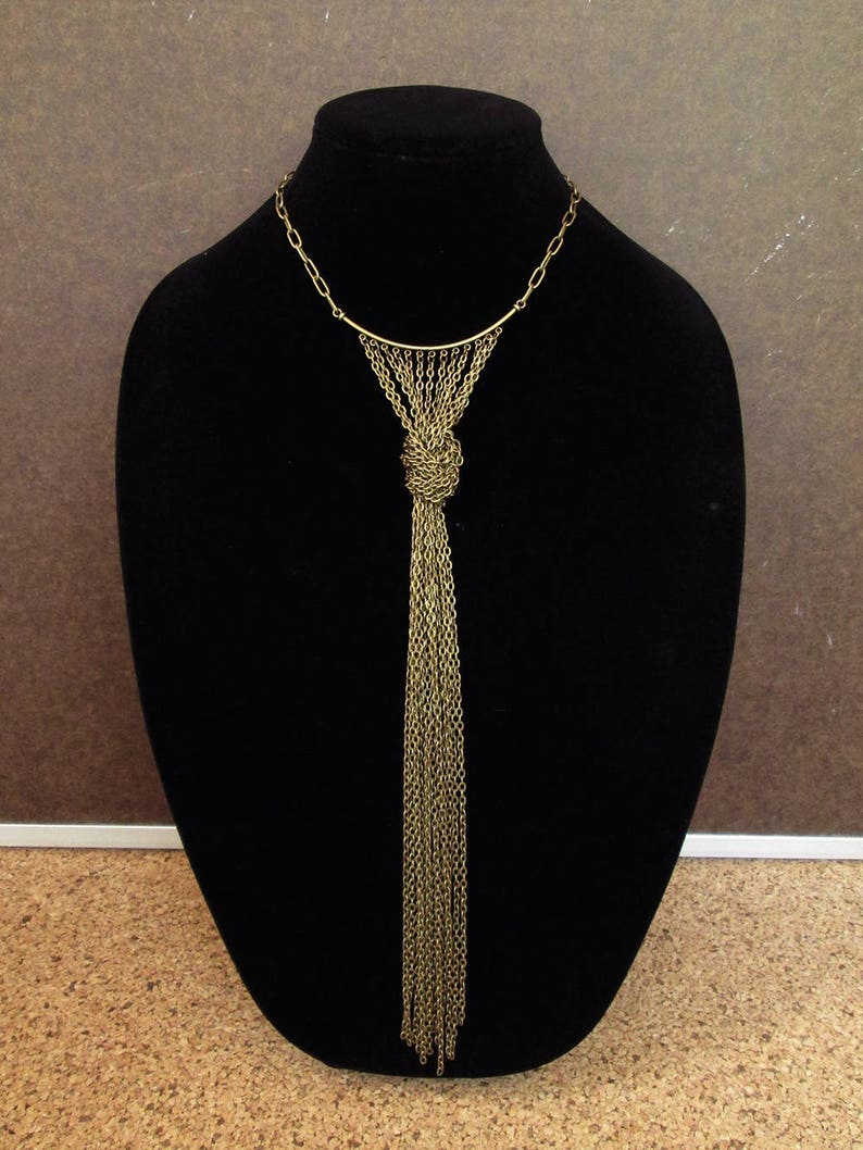 Puede incluir: Un collar de cadena dorada con un largo fleco. El collar tiene una barra simple y curvada en la parte superior y un nudo en la parte inferior donde comienza el fleco.