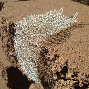 May include: A silver metal mesh fabric with a delicate, open weave. The fabric is draped over a rough, brown surface.