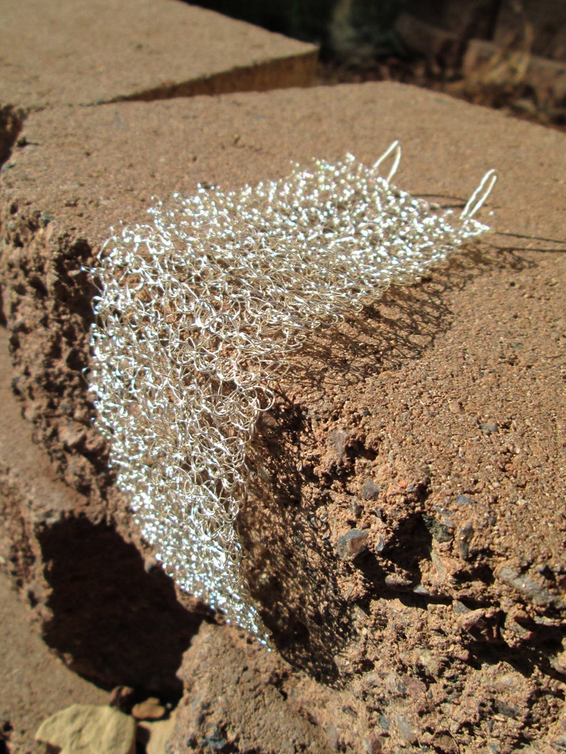 May include: A silver metal mesh fabric with a delicate, open weave. The fabric is draped over a rough, brown surface.