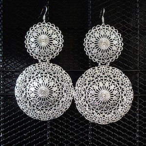 May include: A pair of silver filigree earrings with two circular designs. The earrings are hanging against a black background.