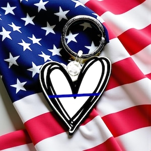 May include: A heart-shaped keychain with a black outline and a blue line across the center, set against a backdrop of the American flag. The keychain has a silver ring for attaching keys.