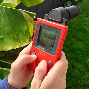 Peut inclure: Une console de jeu vidéo portable Game Boy Pocket rouge avec un accessoire noir. L'écran affiche une scène de jeu. L'appareil est tenu dans les mains, les boutons étant pressés. L'arrière-plan comprend de l'herbe verte et du feuillage.