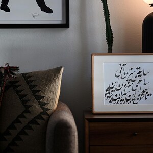 Pu&ograve; includere: Un'opera d'arte incorniciata con calligrafia nera su carta bianca poggia su un comodino di legno. La cornice &egrave; marrone chiaro. Un cuscino fantasia e una silhouette incorniciata sono sullo sfondo. Una lampada nera &egrave; a destra.
