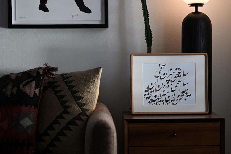 Pu&ograve; includere: Un'opera d'arte incorniciata con calligrafia nera su carta bianca poggia su un comodino di legno. La cornice &egrave; marrone chiaro. Un cuscino fantasia e una silhouette incorniciata sono sullo sfondo. Una lampada nera &egrave; a destra.