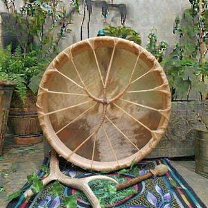 Può includere: Un tamburo a cornice rotondo con una pelle animale marrone chiaro, fissata da sottili lacci in pelle che formano un motivo a stella sul retro. Un martelletto in legno con punta imbottita e un palco di cervo sono posizionati davanti al tamburo.