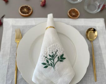 May include: A white linen napkin with an embroidered floral design, folded and held by a gold ring, sits on a white plate. Gold cutlery and a clear glass with a floral pattern are also on the table setting. The table setting includes a white placemat.