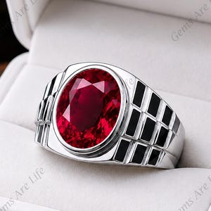Pode incluir: Um anel de tom prateado com uma grande pedra preciosa de cor vermelho-escura e corte oval no centro. A banda é larga e decorada com um padrão de grelha de incrustações retangulares pretas em ambos os lados, apresentado numa caixa branca.