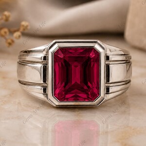 May include: A silver-toned ring featuring a large, rectangular, emerald-cut deep red gemstone set in a polished metal band. The band has a wide, structured design with horizontal grooves on both sides of the central stone setting.