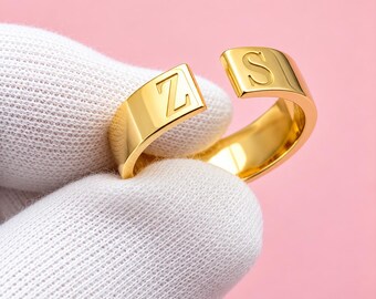 Puede incluir: Un anillo abierto de tono dorado con acabado pulido, que presenta la letra Z grabada en un extremo y la letra S en el otro. La banda tiene una superficie lisa y plana, con un diseño ancho y abierto que permite ajustar su tamaño.