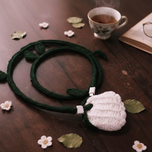 May include: A handmade white crochet earbud case with a dark green crochet cord and leaves. The cord is connected to a white USB-C connector. The scene includes a cup of tea, a book, and decorative flowers and leaves.