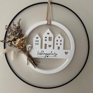 Könnte beinhalten: Weißer Holz-Wandbehang mit drei Haussilhouetten, Herz- und Sternaussparungen und dem Wort "Lieblingsplatz". Das Design befindet sich in einem weißen Kreis, umgeben von einem schwarzen Metallring und mit Trockenblumen verziert.