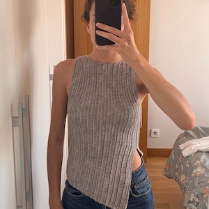 May include: A sleeveless, ribbed knit top in a light brown, heathered yarn. The garment features a high, rounded neckline and an asymmetrical hemline that is shorter on one side, creating a distinct side slit that exposes the waist area.