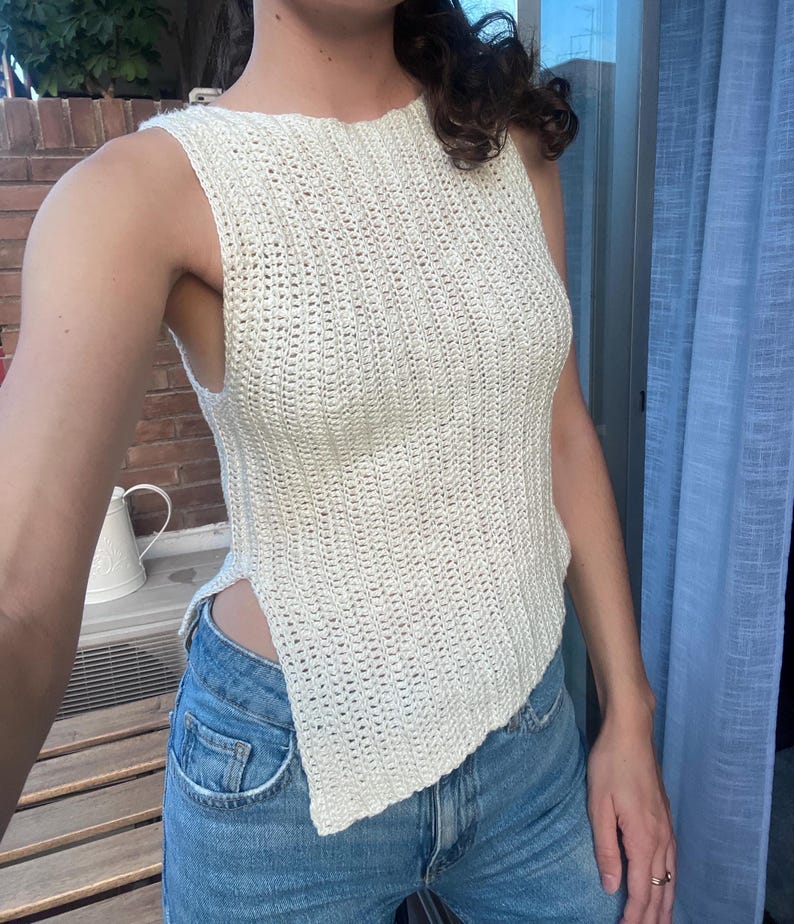 May include: Cream-colored, sleeveless crochet top with a unique asymmetrical hemline. The top has a textured, open weave design. The garment is paired with blue denim jeans. The top is a handmade, bohemian style garment.