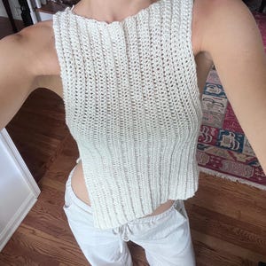 May include: Cream-colored, sleeveless crochet top with a ribbed texture. The top has a high neckline and a slightly cropped length. Paired with light-colored pants. The outfit is casual and appears handmade.