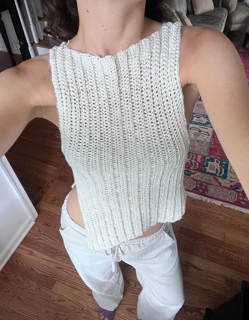 May include: Cream-colored, sleeveless crochet top with a ribbed texture. The top has a high neckline and a slightly cropped length. Paired with light-colored pants. The outfit is casual and appears handmade.