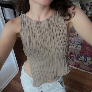May include: A tan crochet top with a unique asymmetrical hemline. The sleeveless top has a ribbed texture and a scoop neckline. The model is also wearing white pants and flower-shaped earrings with red and orange accents.