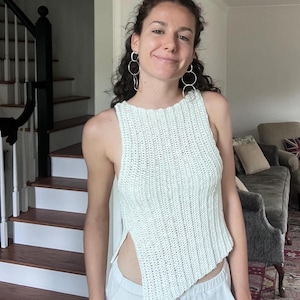 May include: A light green, sleeveless, crochet top with a unique asymmetrical hemline. The top is paired with white pants. The image is taken indoors, with a staircase and a living room visible in the background.