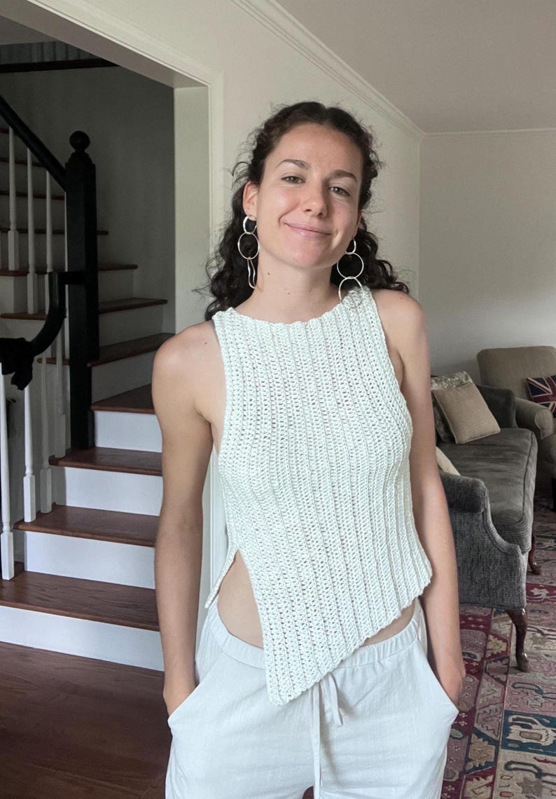 May include: A light green, sleeveless, crochet top with a unique asymmetrical hemline. The top is paired with white pants. The image is taken indoors, with a staircase and a living room visible in the background.