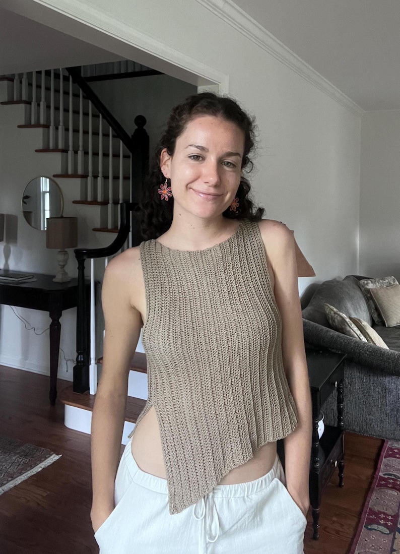 May include: A tan, sleeveless, ribbed knit top with an asymmetrical hemline. The top is paired with white pants. The person is wearing flower-shaped earrings.