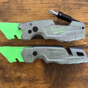 May include: Two gray and green utility knives with a topographic pattern. The knives have bright green blades and the text "Milwaukee FASTBACK". One knife has a black screwdriver bit attached.