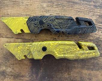 May include: Two Milwaukee Fastback utility knives featuring a topographic map pattern. The top knife is black with yellow lines, and the bottom knife is yellow with black lines. Both knives have a folding design, a belt clip, and a blade release button.