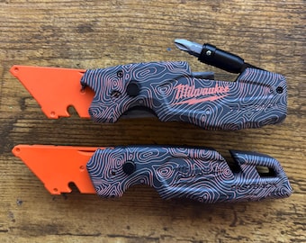 May include: Two Milwaukee Fastback utility knives with orange blades and black handles featuring a topographic line pattern. The top knife has an extended screwdriver bit, while the bottom knife shows the Fastback branding on the handle.