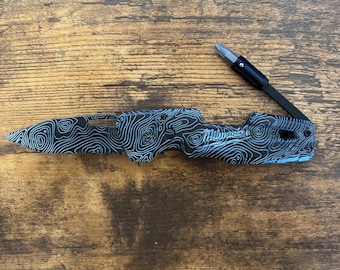 May include: A black and blue folding knife with a topographic pattern. The knife has a partially serrated blade and a black handle. The text "Milwaukee" is visible on the handle. The knife is open, revealing a screwdriver bit.