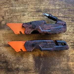 May include: Two Milwaukee Fastback utility knives with orange blades and black handles featuring a copper-coloured topographic line pattern. The top knife includes an extended screwdriver bit holder, while the bottom knife shows the integrated blade release.