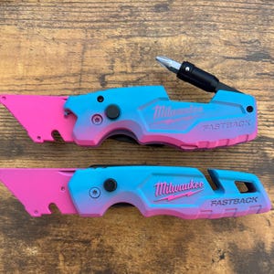 May include: Two Milwaukee Fastback utility knives featuring a vibrant blue and pink gradient finish. The top knife includes an extended screwdriver bit, while both display the Milwaukee logo and Fastback text in a bold, embossed design.