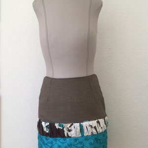 Pode incluir: Uma saia pelo joelho exibida num manequim. A parte superior apresenta um padr&atilde;o de micro-xadrez castanho e preto. Abaixo, h&aacute; um folho franzido com estampado floral castanho, branco e turquesa, seguido de um painel turquesa com bordados.