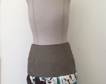 Puede incluir: Una falda hasta la rodilla sobre un maniquí. La parte superior presenta un patrón de microcuadros marrones y negros. Debajo hay un volante fruncido con estampado floral marrón, blanco y turquesa, seguido de un panel turquesa con bordados negros.