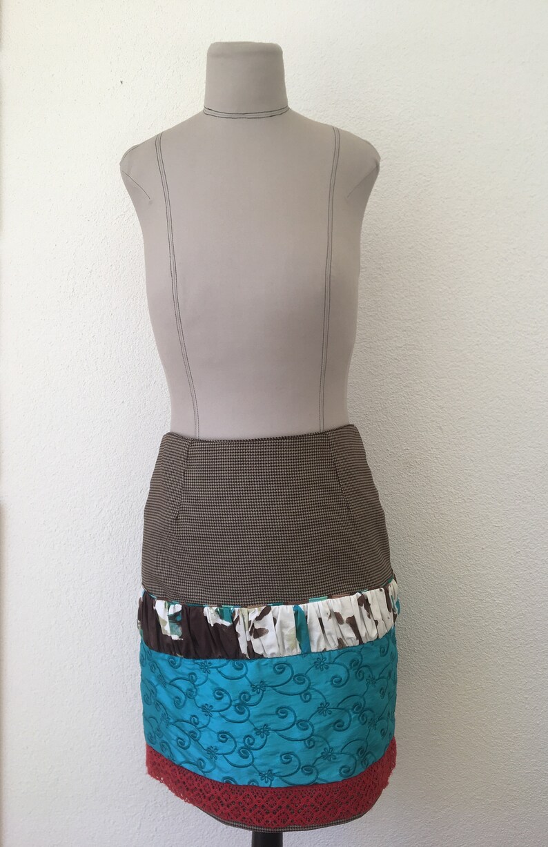 Pode incluir: Uma saia pelo joelho exibida num manequim. A parte superior apresenta um padr&atilde;o de micro-xadrez castanho e preto. Abaixo, h&aacute; um folho franzido com estampado floral castanho, branco e turquesa, seguido de um painel turquesa com bordados.