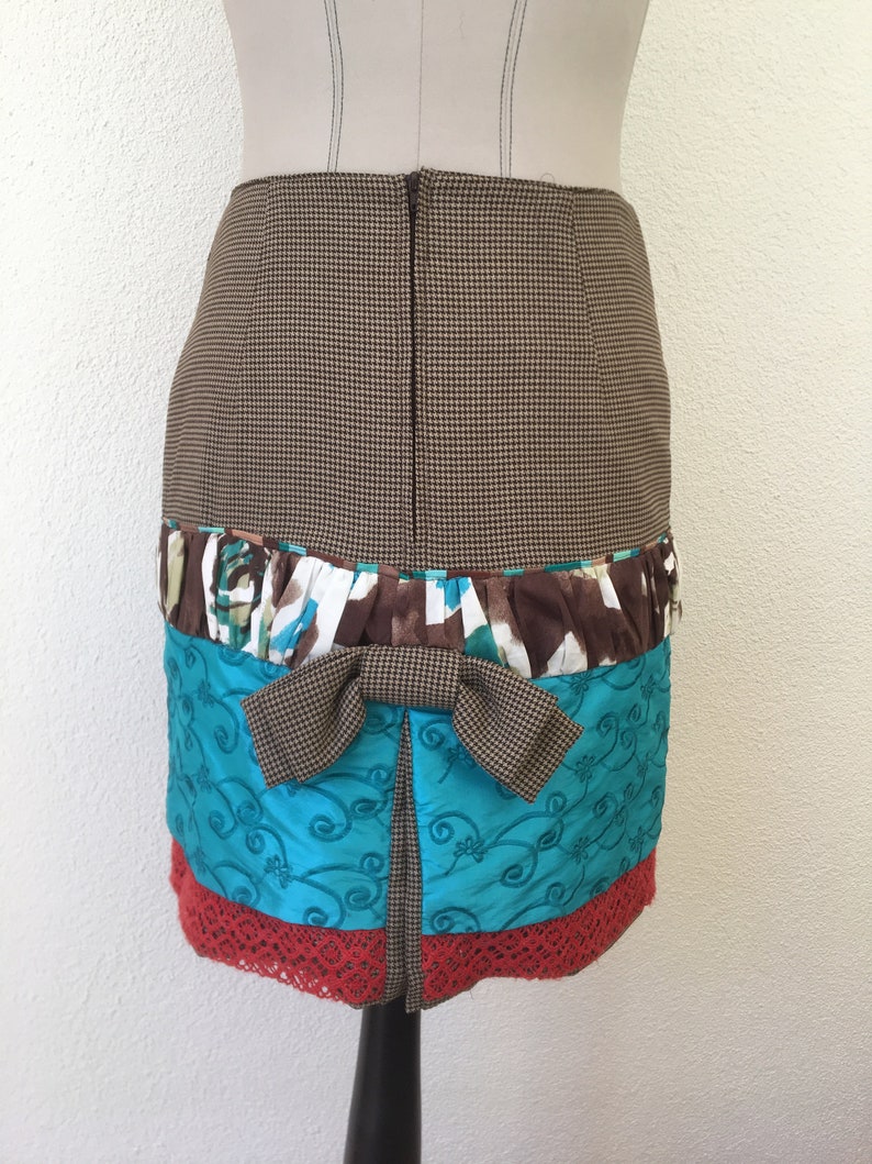 Pode incluir: Uma saia pelo joelho com um padr&atilde;o de p&eacute;-de-galinha castanho e bege na parte superior, um la&ccedil;o central de p&eacute;-de-galinha castanho, uma faixa com folhos em estampado floral castanho, branco e turquesa, um painel bordado turquesa e uma bainha de renda vermelha.