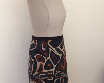 Pode incluir: Uma saia até o joelho com um padrão geométrico abstrato arrojado em tons de azul escuro, laranja queimado, creme e preto. A saia tem um cós preto sólido e é exibida num manequim de cor neutra para mostrar o ajuste da peça.