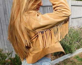 May include: A tan suede jacket with fringe detailing on the sleeves. The jacket is paired with light blue denim shorts with distressed detailing. The image is taken outdoors with a wooden fence and greenery in the background.