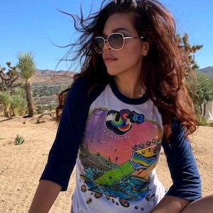 May include: A person wearing a white and navy blue raglan sleeve t-shirt with a colorful graphic design. The graphic features a guitar and the text "Festival '83". The person is also wearing sunglasses.