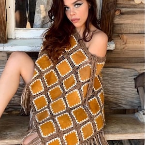 May include: A brown and tan poncho with a square pattern and fringe. The poncho is made of a woven material with tan squares and brown borders. The fringe is tan and white.
