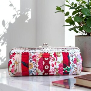 May include: A rectangular clutch purse featuring a patchwork design of various floral and solid red fabric strips. The purse has a silver-toned metal kiss-lock frame, a white lace trim along the top edge, and a small red oval brand label on the front.