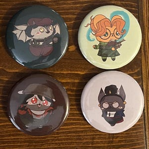 Peut inclure: Quatre badges ronds avec des illustrations de personnages de dessin animé. En haut à gauche, un gris foncé avec une créature ailée, en haut à droite, un vert clair avec un personnage aux cheveux orange, en bas à gauche, un marron, et en bas à droite, un violet.