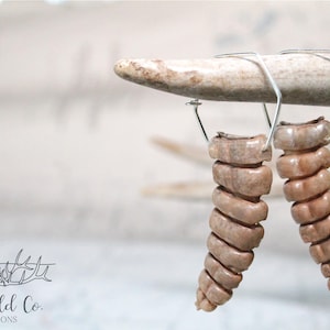 May include: A pair of spiral-shaped earrings with a light brown color, suspended from silver-colored wire hooks. The earrings are displayed against a blurred background and a light-colored antler. The logo "Lw Wild Co. Creations" is in the lower left corner.