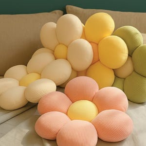 Puede incluir: Primer plano de varias almohadas con forma de flor en varios colores. Las almohadas tienen un aspecto suave y texturizado con un diseño acanalado. Los colores incluyen amarillo pálido, rosa claro y verde. Las almohadas están dispuestas sobre una superficie gris claro.